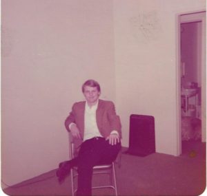 Here's an alt tag for the image: Smiling man sitting in chair, 1970s photo.